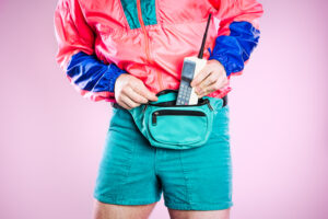 A man wearing fluorescent colored clothing puts a 1980's - 1990's cellular brick phone into his fanny pack, representing state of the art style and technology for that time. Detail shot; horizontal with pink background.